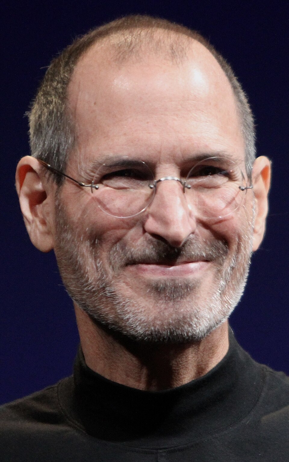 Steve Jobs, smiling, in a black turtleneck and rimless glasses.